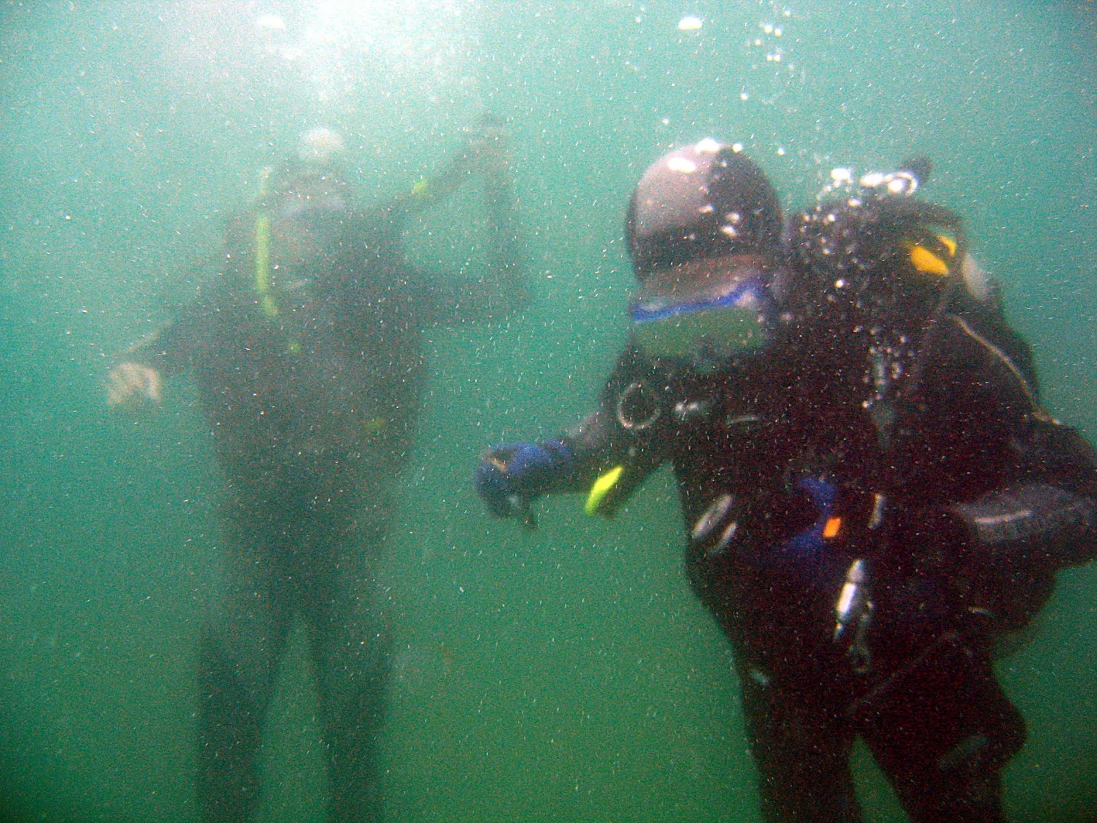 Zwei Taucher in dunklen Neoprenanzügen unter Wasser, im Vordergrund mit Atemregler und Tarierweste, Luftblasen steigen auf, grünblaues trübes Wasser.