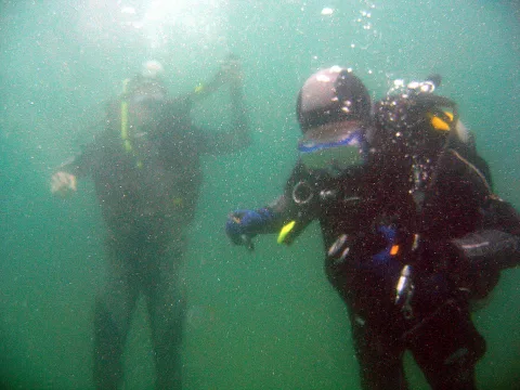 Zwei Taucher in dunklen Neoprenanzügen unter Wasser, im Vordergrund mit Atemregler und Tarierweste, Luftblasen steigen auf, grünblaues trübes Wasser.