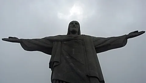 Christusstatue mit ausgebreiteten Armen vor grauem, bewölktem Himmel, von unten fotografiert