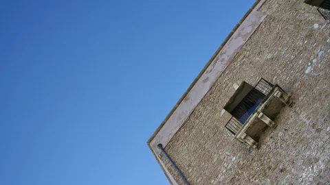 Ecke eines Steinhauses mit vergittertem Fenster und steinernem Balkon vor blauem Himmel, von unten fotografiert.
