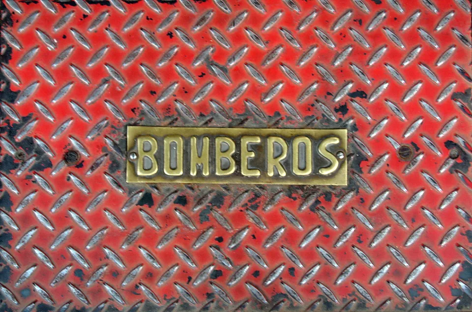 Ein rechteckiges Messingschild mit der Aufschrift BOMBEROS in erhabenen Buchstaben sitzt mittig auf einer rot lackierten Riffelblechfläche mit abgeblätterter Farbe.
