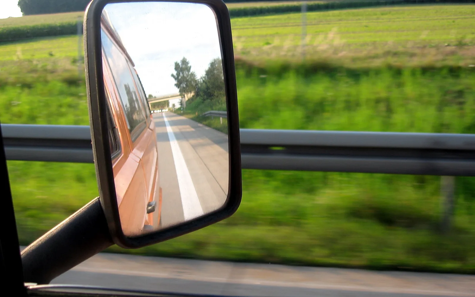 Rechteckiger Seitenspiegel eines Autos, in dem eine Straße und ein orangefarbener Wagen zu sehen sind, davor verschwommene grüne Wiese und Leitplanke.