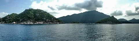 Panoramablick vom Boot auf das ruhige Meer mit bewaldeten Felsinseln unter teils bewölktem Himmel