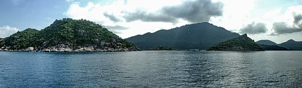 Panoramablick vom Boot auf das ruhige Meer mit bewaldeten Felsinseln unter teils bewölktem Himmel