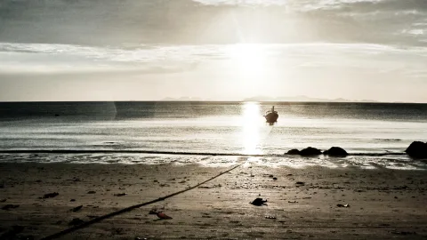 Ein kleines Boot liegt im flachen Wasser vor einem Sandstrand, im Gegenlicht der tief stehenden Sonne mit Wolkenband am Himmel.