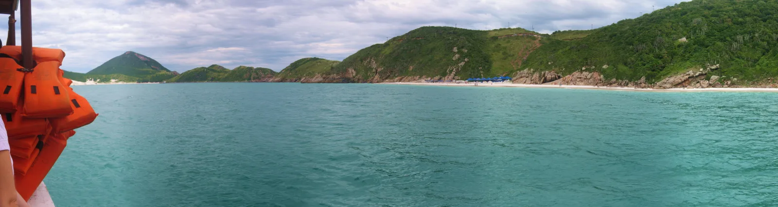 Panoramablick von einem Boot auf eine türkisfarbene Bucht, links eine Reihe orangefarbener Schwimmwesten, dahinter bewaldete Hügel und ein heller Sandstrand mit blauem Pavillon.
