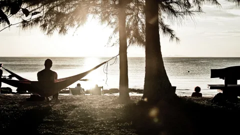 Ein Mann sitzt in einer Hängematte zwischen Palmen am Strand, im Gegenlicht der tiefstehenden Sonne, dahinter liegt ruhiges Meer.