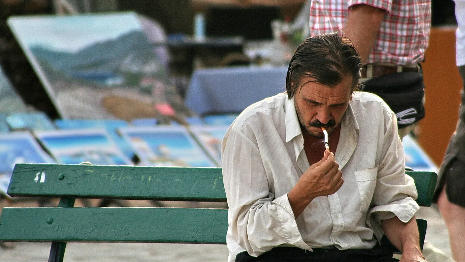 Ein Mann mit zurückgekämmtem dunklem Haar und Schnurrbart sitzt auf einer grünen Parkbank und hält eine Zigarette an die Lippen, im Hintergrund stehen Gemälde auf einem Markt.