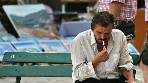 Ein Mann mit zurückgekämmtem dunklem Haar und Schnurrbart sitzt auf einer grünen Parkbank und hält eine Zigarette an die Lippen, im Hintergrund stehen Gemälde auf einem Markt.