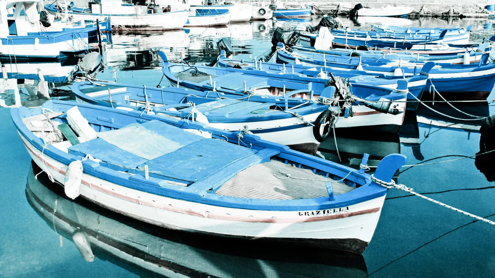 Mehrere blau-weiße Fischerboote liegen dicht nebeneinander in einem ruhigen Hafenbecken, im Vordergrund ist ein Boot mit der Aufschrift Graziella zu sehen, dessen Spiegelung im Wasser sichtbar ist.