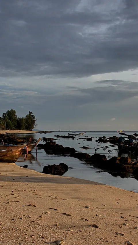 Sandstrand im Vordergrund, links ein holzernes Langheckboot am Ufer, dahinter flaches Wasser mit dunklen Felsen, Bäume am linken Rand und bewölkter Himmel.