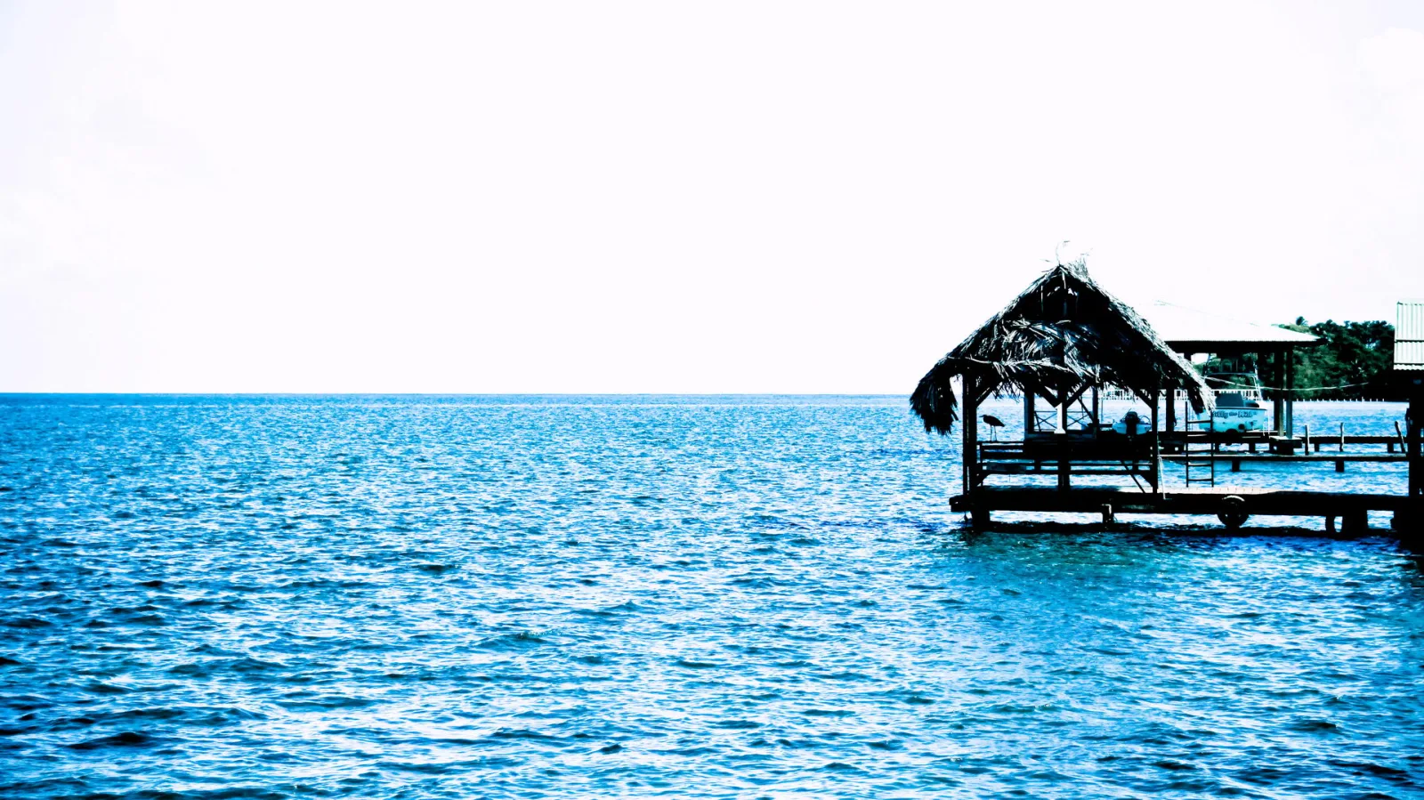 Eine Holzhütte mit Strohdach steht auf einem Steg über dem blauen Meer, links ist nur Wasser und heller Himmel zu sehen.