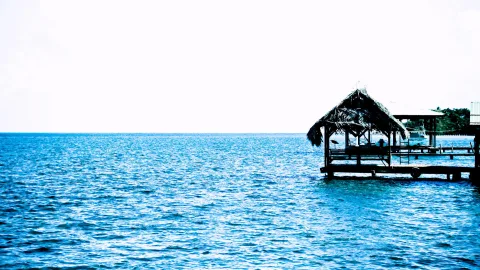Eine Holzhütte mit Strohdach steht auf einem Steg über dem blauen Meer, links ist nur Wasser und heller Himmel zu sehen.