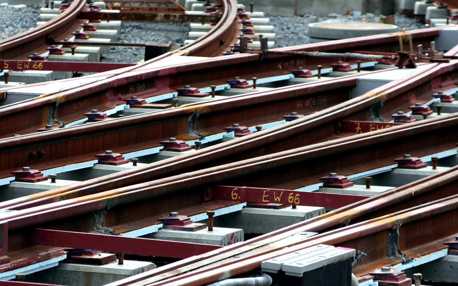 Mehrere rostige Schienen laufen diagonal in einer Gleisweiche zusammen, auf grauen Betonschwellen mit roten Befestigungsplatten und Schrauben, dazu gelbe Kennzeichnungen auf den Schienen.