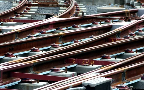 Mehrere rostige Schienen laufen diagonal in einer Gleisweiche zusammen, auf grauen Betonschwellen mit roten Befestigungsplatten und Schrauben, dazu gelbe Kennzeichnungen auf den Schienen.