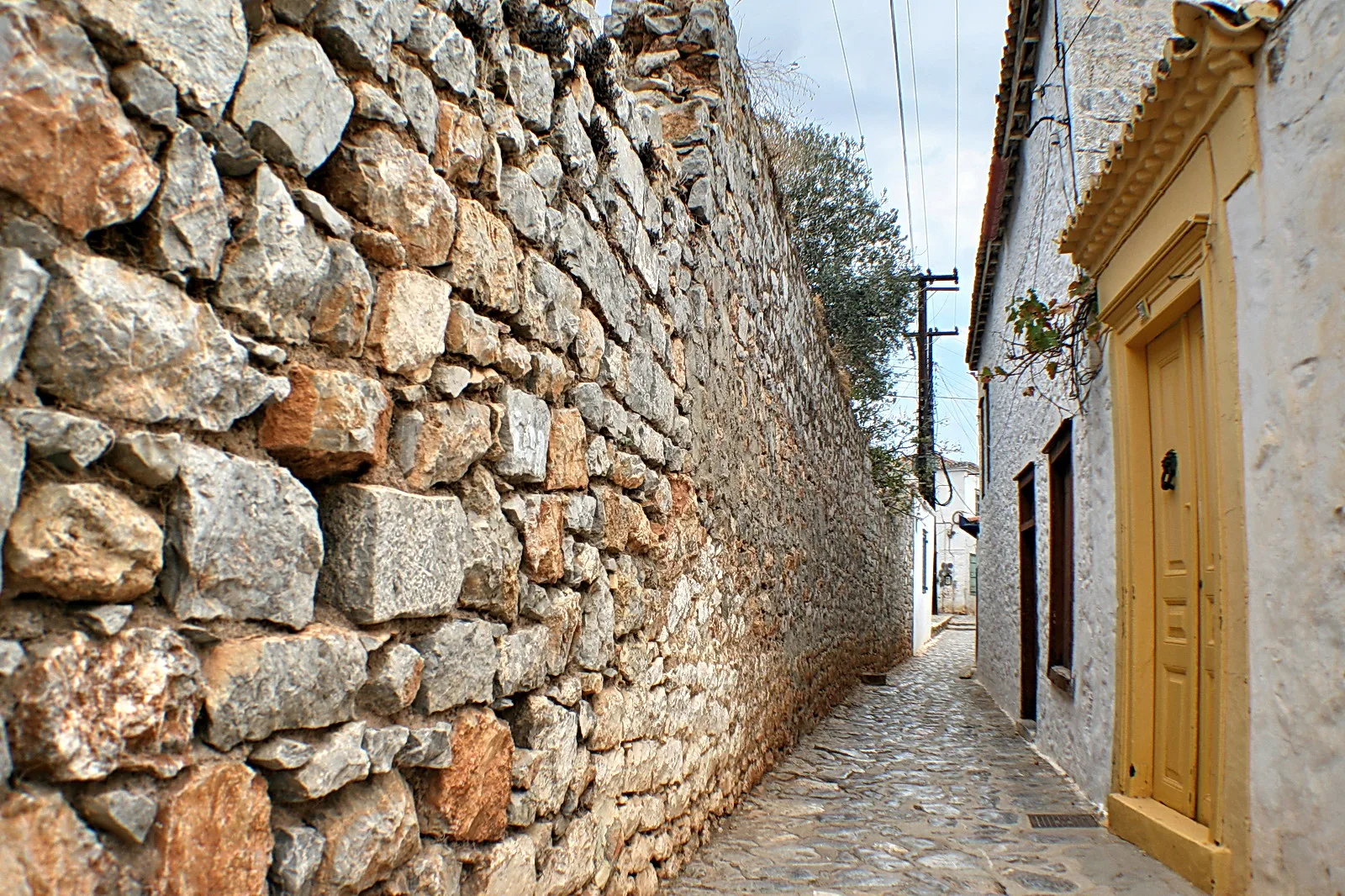 Enge Gasse mit hoher Bruchsteinmauer links, weiß getünchtem Haus mit gelber Tür rechts und Kopfsteinpflaster am Boden.