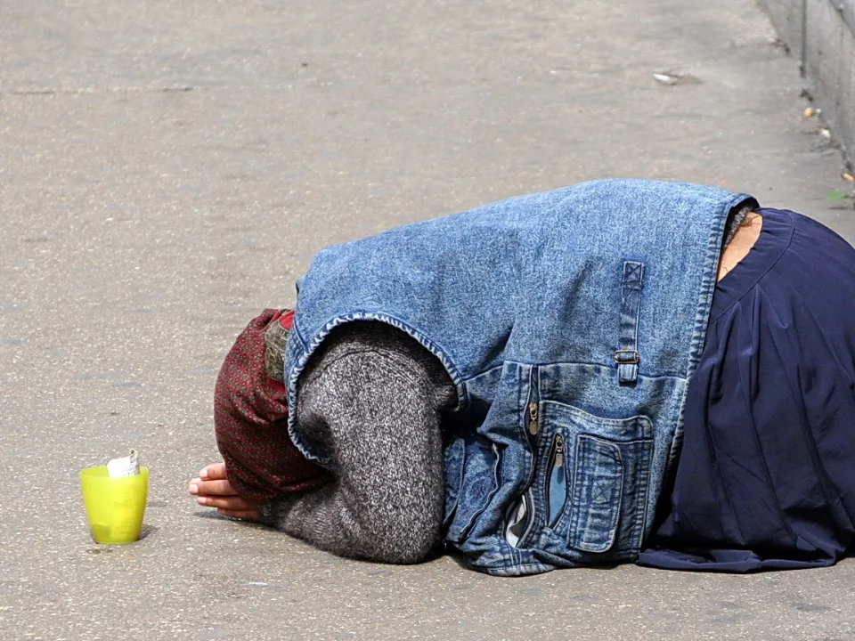 Eine Person kniet auf einem grauen Gehweg, der Kopf ist unter einer blauen Jeansjacke verborgen, daneben steht ein gelber Becher.