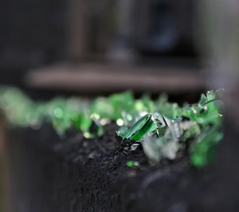 Grüne Glasscherben liegen auf einer dunklen, rauen Mauerkante, der Hintergrund ist unscharf.