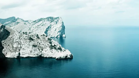 Weiß-graue Felsklippen ragen links ins Bild, daneben liegt eine weite, ruhige blaue Meeresfläche unter bewölktem Himmel.
