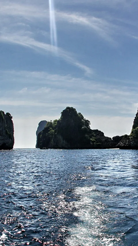 Mehrere bewachsene Felsinseln ragen aus dem Meer, darüber ein blauer Himmel mit weißen Schlieren und einem senkrechten Kondensstreifen.