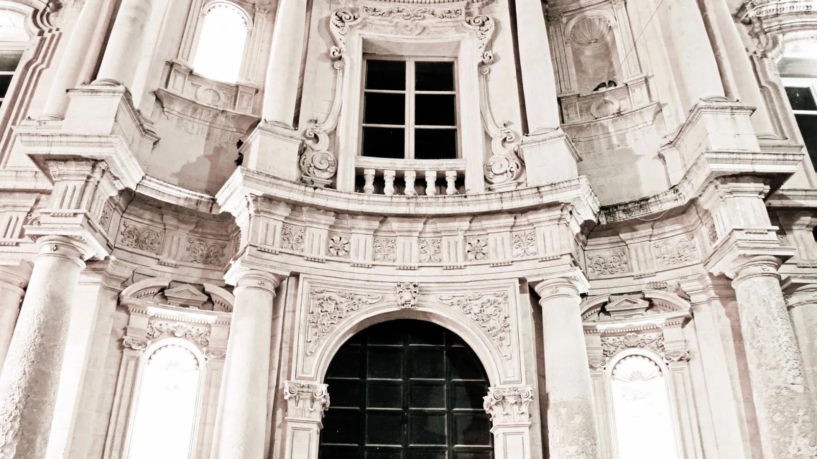 Barocke Steinfassade mit Säulen, Ornamenten und einem Balkon mit Fenster, im unteren Bereich ein dunkles, vergittertes Portal mit Rundbogen.
