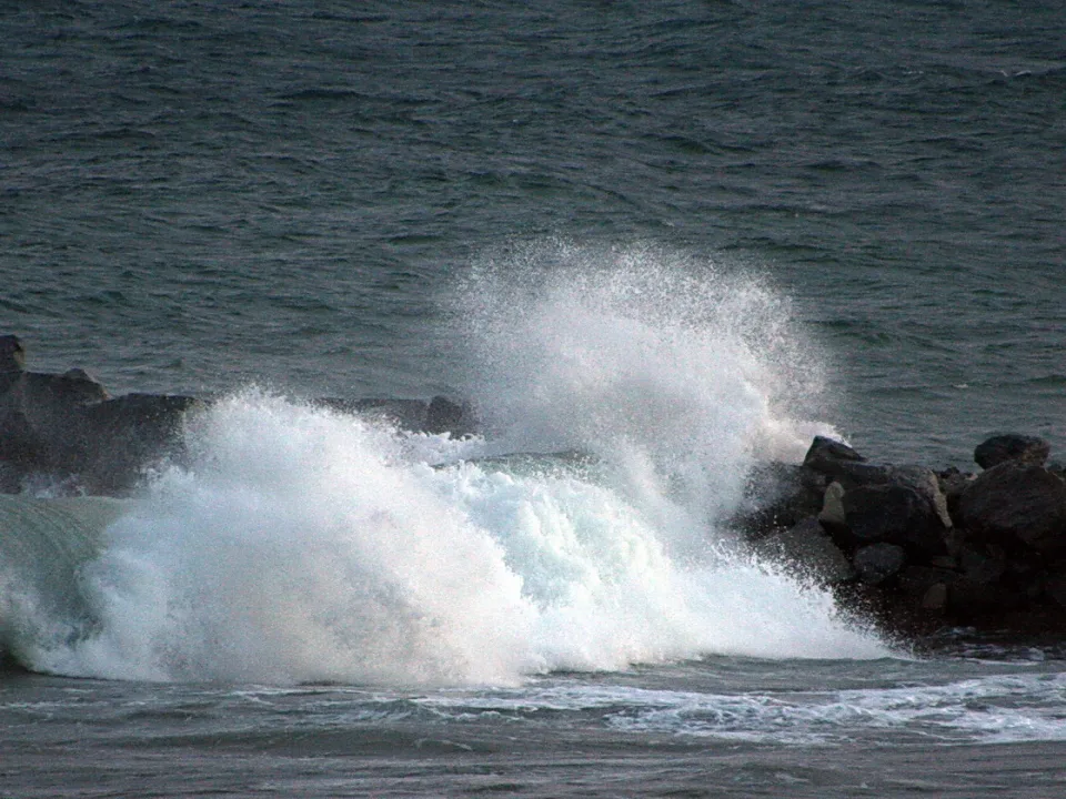 Eine weiße Welle schlägt mit Gischt gegen dunkle Felsen eines Wellenbrechers, dahinter liegt graues Meer.