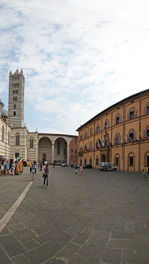 Platz mit Kathedrale aus hellem und dunklem Marmor und gestreiftem Glockenturm, rechts ein orangefarbener Palast mit Fahnen und Fenstern, davor Touristen auf Pflastersteinen