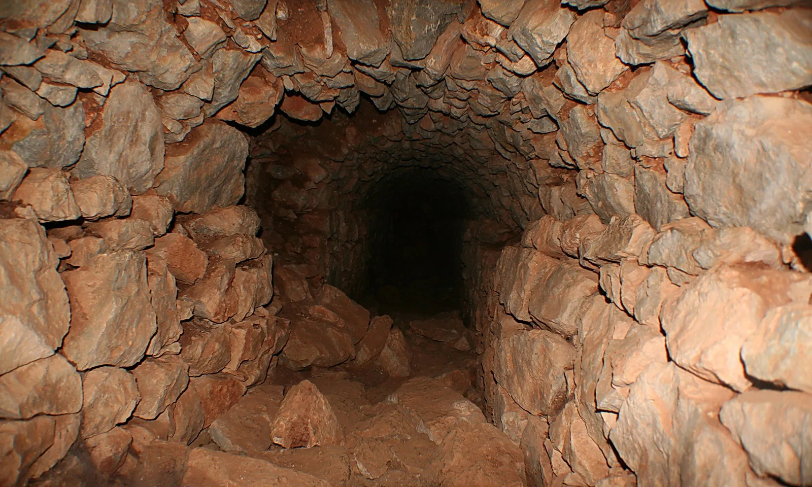 Enger Gang mit unregelmäßig geschichteten rotbraunen Steinwänden, der nach hinten in völliger Dunkelheit verschwindet.
