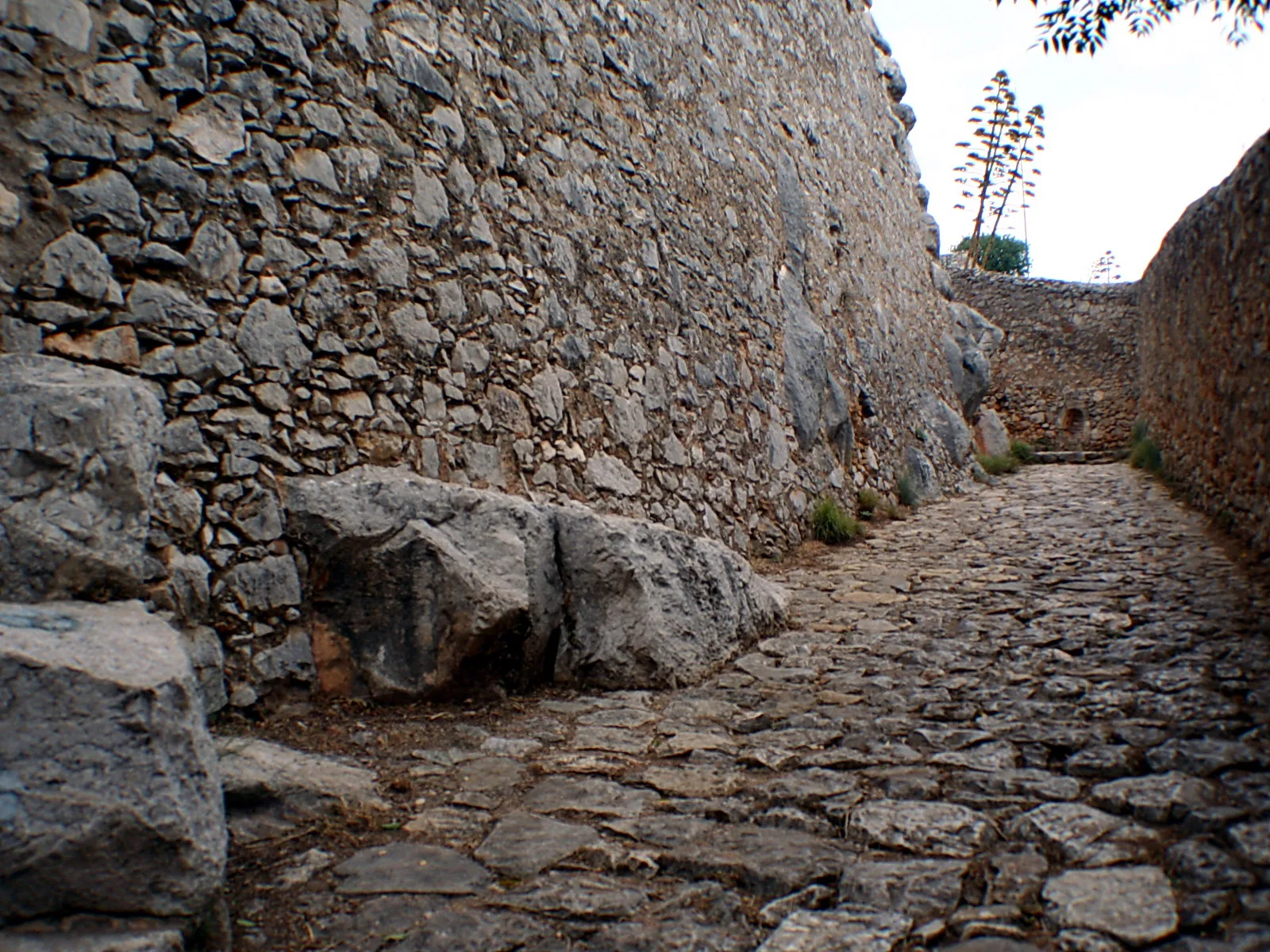 Schmale Gasse mit Kopfsteinpflaster, links eine hohe Bruchsteinmauer mit großen Felsbrocken, rechts eine weitere Mauer, im Hintergrund eine Agave vor hellem Himmel.