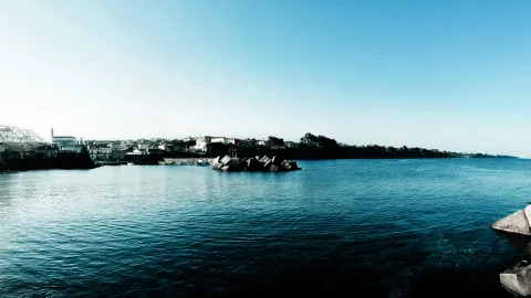 Breites Panorama einer Küstenstadt mit Häusern und einem Kirchturm am Ufer, davor ruhiges blaues Meer und große Betonblöcke im Wasser, darüber klarer blauer Himmel.
