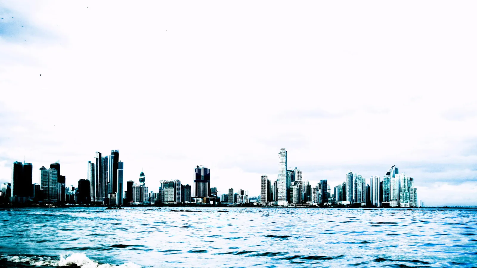 Skyline mit dicht stehenden Hochhäusern hinter einer breiten Wasserfläche unter hellem, bewölktem Himmel.