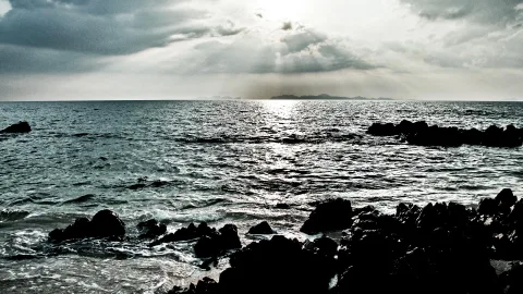 Dunkle Felsen im Vordergrund, dahinter ein unruhiges Meer unter bewölktem Himmel, die Sonne bricht durch die Wolken und spiegelt sich auf dem Wasser. Am Horizont liegen flache Inselsilhouetten.