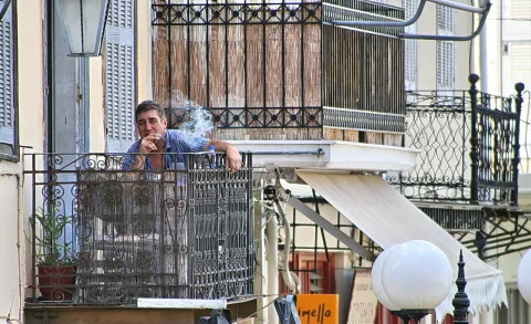 Ein Mann mit kurzem dunklem Haar und kariertem Hemd lehnt rauchend am schmiedeeisernen Balkongeländer eines alten Hauses, links steht ein Blumentopf mit Pflanze.