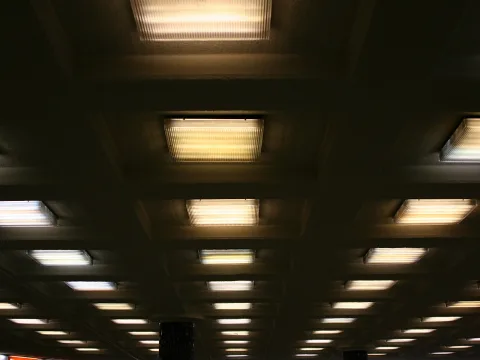 Decke mit einem Raster aus eckigen Deckenleuchten mit geriffeltem Glas, die warm leuchten; im Vordergrund ein dunkler Pfeiler.
