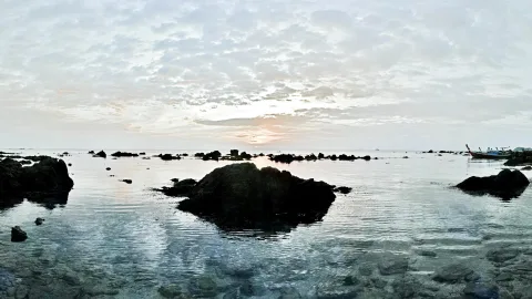 Dunkle Felsen ragen aus ruhigem, flachem Meerwasser. Im Hintergrund hängt eine bewölkte Dämmerung über dem Horizont, rechts liegt ein kleines Boot.