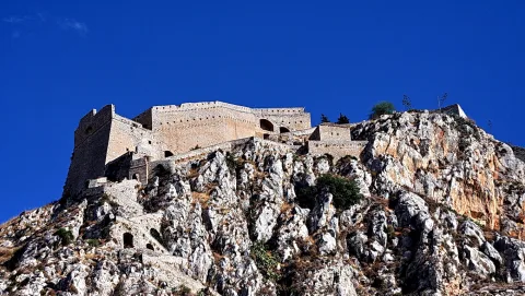 Eine alte Festung mit hellen Mauern, Bogenöffnungen und Zinnen thront auf einem zerklüfteten Felsen unter tiefblauem Himmel.