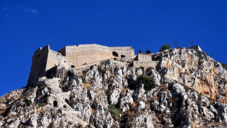 Eine alte Festung mit hellen Mauern, Bogenöffnungen und Zinnen thront auf einem zerklüfteten Felsen unter tiefblauem Himmel.