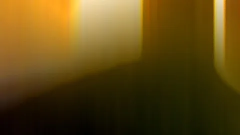 Abstrakte, verwischte Aufnahme mit orangefarbenem und gelbem Licht oben links, helle Streifen in der Mitte und ein dunkler, olivgrüner Schatten im unteren Bereich.