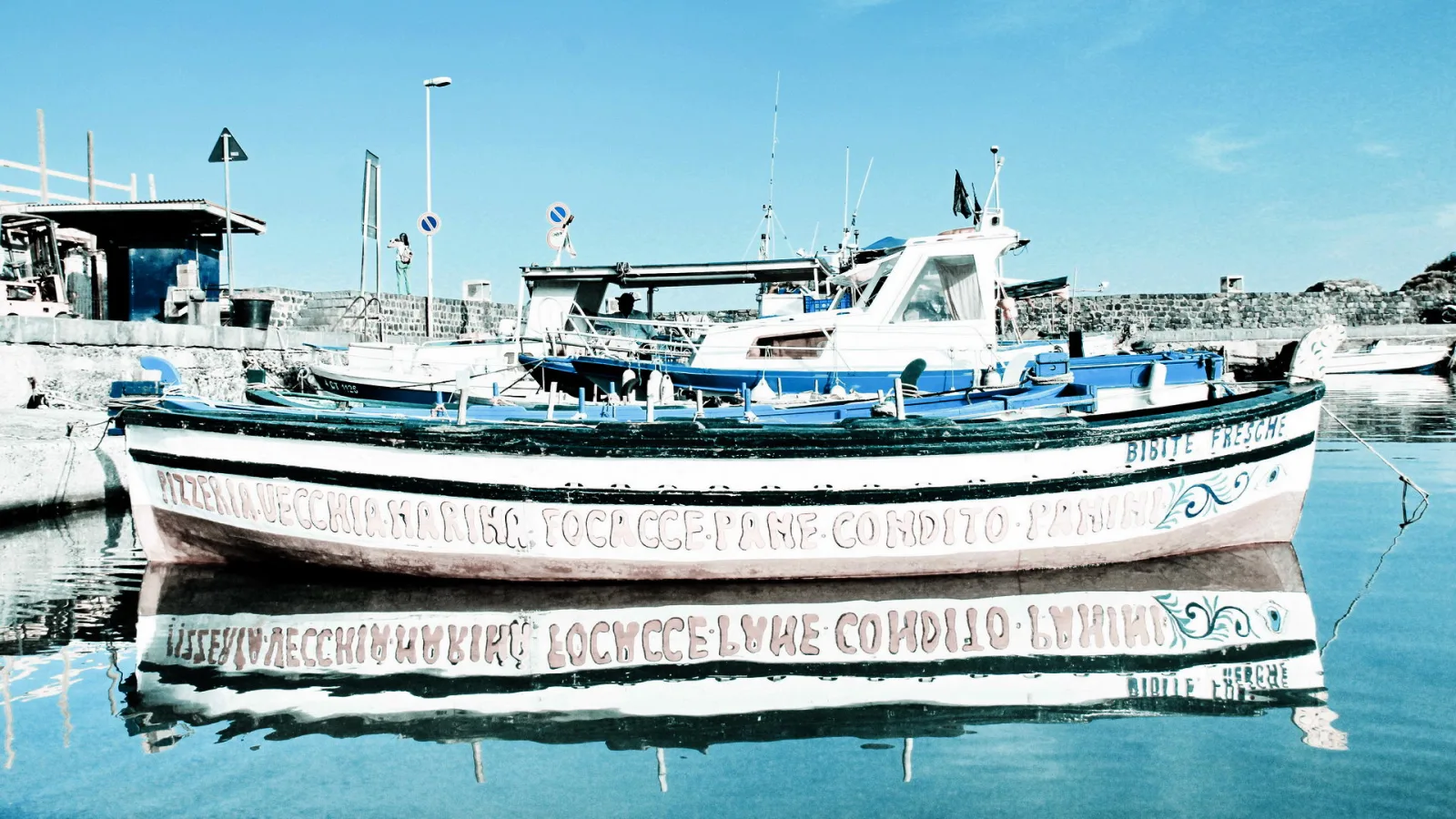 Weiß und blau lackiertes Fischerboot mit beschrifteter Bordwand liegt im ruhigen Hafenbecken, das Boot spiegelt sich im Wasser.
