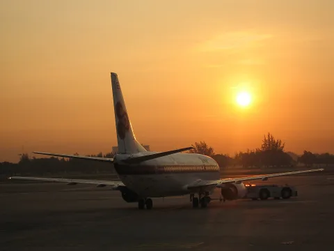 Ein Passagierflugzeug rollt im Gegenlicht über das Vorfeld, dahinter steht die tief stehende Sonne vor orangefarbenem Himmel.