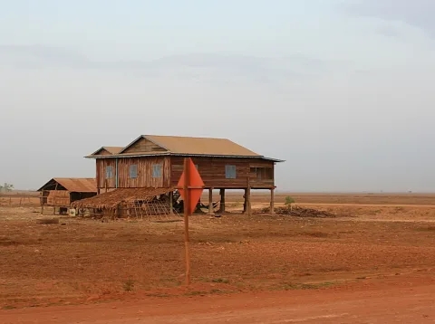 Holzhaus auf hohen Stelzen mit Wellblechdach in einer trockenen, rötlich braunen Ebene, links eine kleine Hütte, vorne ein orangefarbener Pfahl