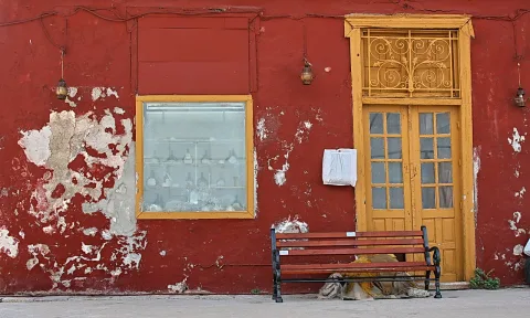 Rot verputzte, abblätternde Hauswand mit gelber Holztür, vergittertem Oberlicht, Schaufenster und einer Holzbank davor.