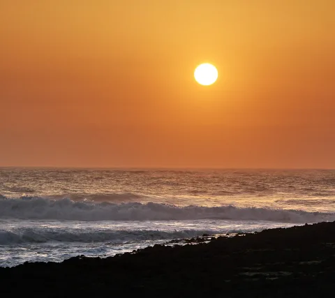 Große orangefarbene Sonne über einem weiten Meer, im Vordergrund brechen Wellen an einem dunklen Felsstrand.