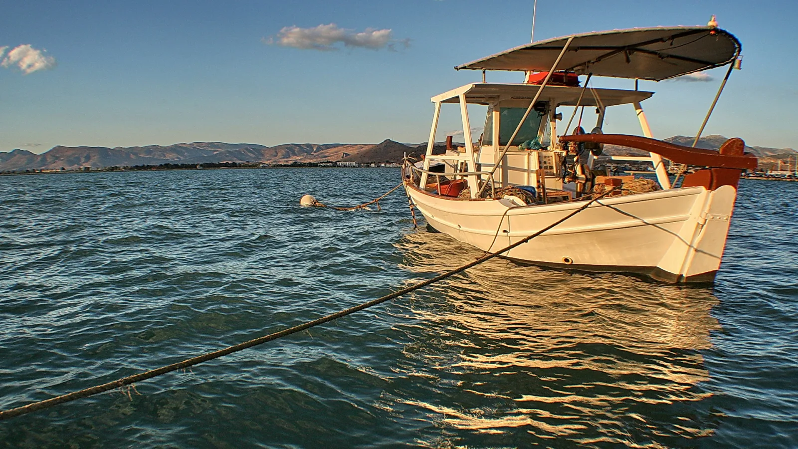 Ein weißes Fischerboot mit rotbraunem Bug und Sonnendach liegt auf ruhigem, türkisfarbenem Wasser vor einer Küste mit Bergen am Horizont. Ein Seil führt diagonal aus dem Bild, eine weiße Boje treibt links im Wasser.