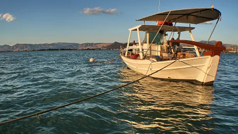 Ein weißes Fischerboot mit rotbraunem Bug und Sonnendach liegt auf ruhigem, türkisfarbenem Wasser vor einer Küste mit Bergen am Horizont. Ein Seil führt diagonal aus dem Bild, eine weiße Boje treibt links im Wasser.