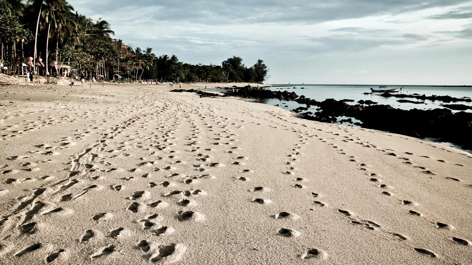 Sandstrand mit vielen Fußspuren, links Palmen und Bäume, rechts dunkle Felsen im flachen Meer, im Hintergrund ein Boot am Horizont unter bewölktem Himmel.