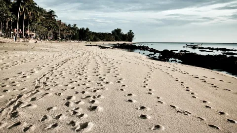 Sandstrand mit vielen Fußspuren, links Palmen und Bäume, rechts dunkle Felsen im flachen Meer, im Hintergrund ein Boot am Horizont unter bewölktem Himmel.