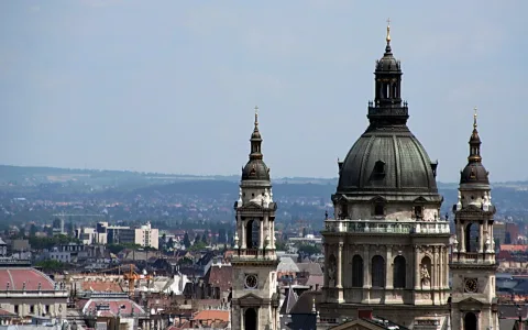 Die Kuppel und zwei Türme der Szent-István-Basilika ragen über den Dächern von Budapest auf, dahinter liegen Hügel im Dunst unter blassblauem Himmel.