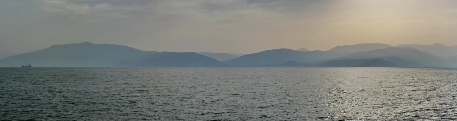 Breite Meeresfläche mit leichten Wellen unter diesigem Himmel, dahinter ein Bergzug im Dunst, links am Horizont ein kleines Schiff.