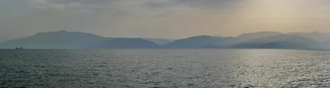 Breite Meeresfläche mit leichten Wellen unter diesigem Himmel, dahinter ein Bergzug im Dunst, links am Horizont ein kleines Schiff.